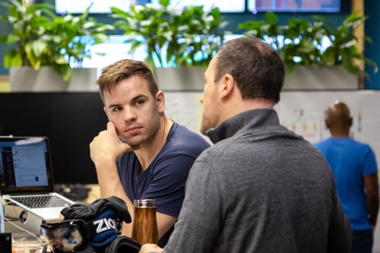 Two men in casual attire have discussion at an office desk.